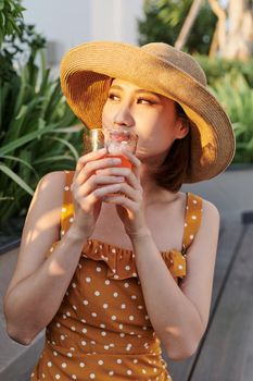 Beautiful Asian girl siting in the bench and drinking juice under summer sunshine.