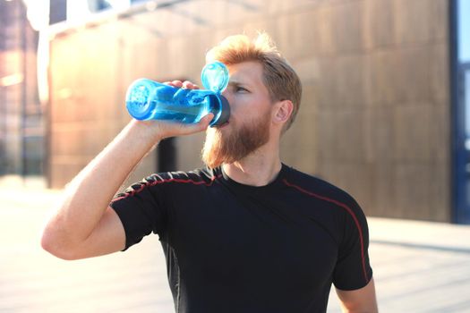 Handsome adult man drinking water from fitness bottle while standing outside, at sunset or sunrise. Runner