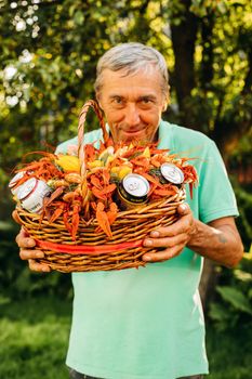 Portrait of an old caucasian grandfather with fair hair in t-shirt holds a large basket with cooked red large crays, smiling and enjoying of his life