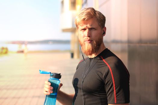 Handsome adult man drinking water from fitness bottle while standing outside, at sunset or sunrise. Runner