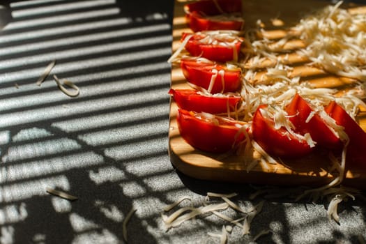 Stock photo in close up of sliced tomatoes with shredded cheese on wooden board in sunlight. Blinds shadow over the table. Cooking process.