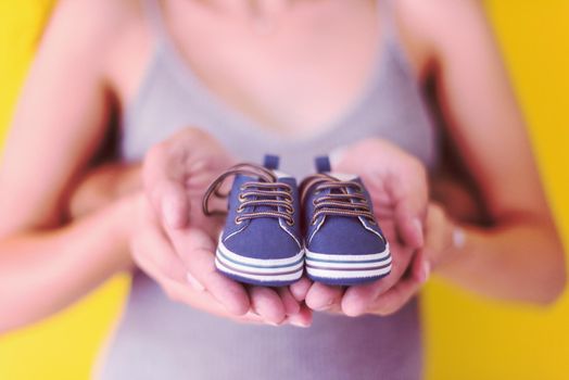 young  pregnant couple holding newborn baby shoes isolated on yellow background,family and parenthood concept