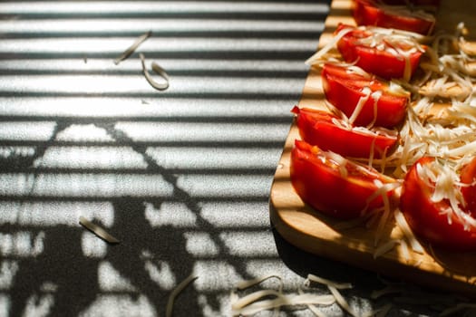 Stock photo in close up of sliced tomatoes with shredded cheese on wooden board in sunlight. Blinds shadow over the table. Cooking process.