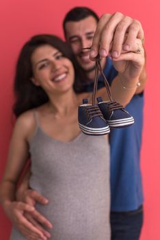 young  pregnant couple holding newborn baby shoes isolated on red background,family and parenthood concept