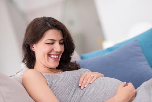 young happy pregnant woman keeping hand on belly while sitting on sofa at home