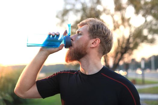 Handsome adult man drinking water from fitness bottle while standing outside, at sunset or sunrise. Runner