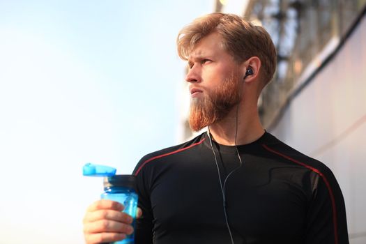 Handsome adult man drinking water from fitness bottle while standing outside, at sunset or sunrise. Runner