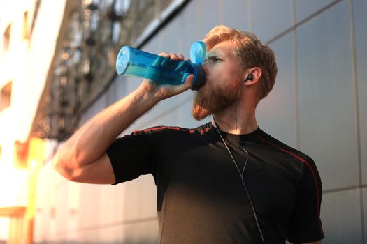 Handsome adult man drinking water from fitness bottle while standing outside, at sunset or sunrise. Runner