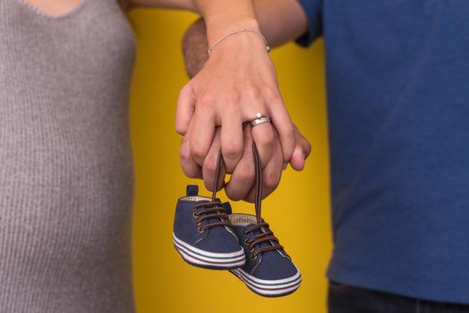 young  pregnant couple holding newborn baby shoes isolated on yellow background,family and parenthood concept