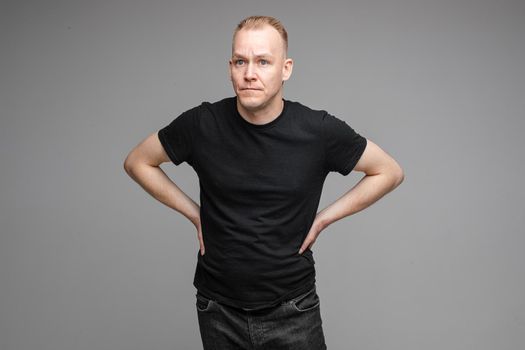 caucasian male wearing a black t-shirt and jeans posing for the camera with hands on a belt on grey background