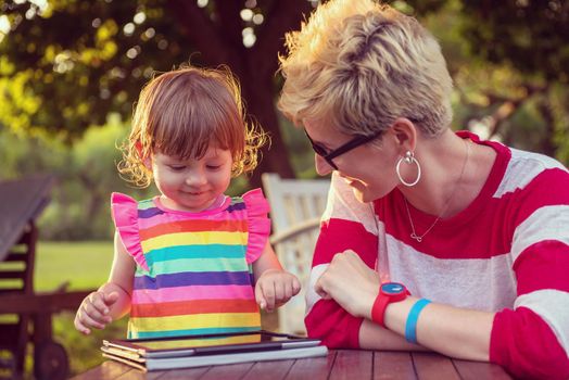 Happy mother and her little daughter enjoying free time using tablet computer while relaxing  on holiday home garden during sunny day