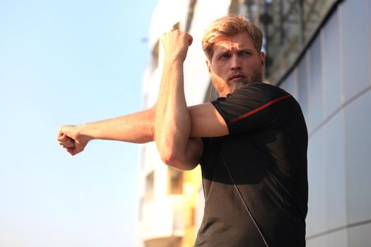 Handsome young man doing stretching exercises before running while standing outdoors