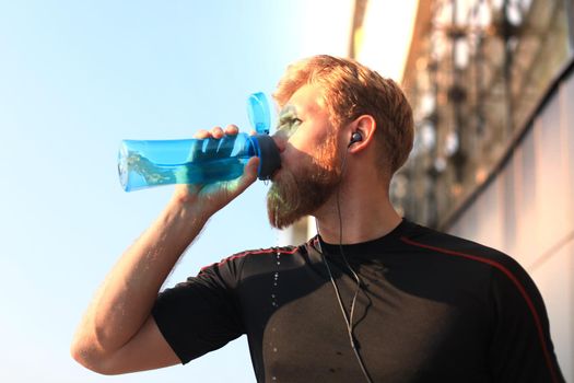 Handsome adult man drinking water from fitness bottle while standing outside, at sunset or sunrise. Runner