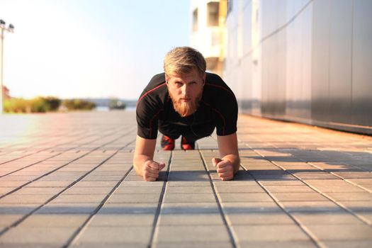 Handsome young man in sports clothing keeping plank position while exercising outdoors