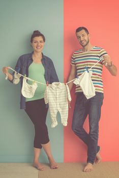 Beautiful pregnant woman and her husband expecting baby holding baby bodysuits and smiling over colorful background