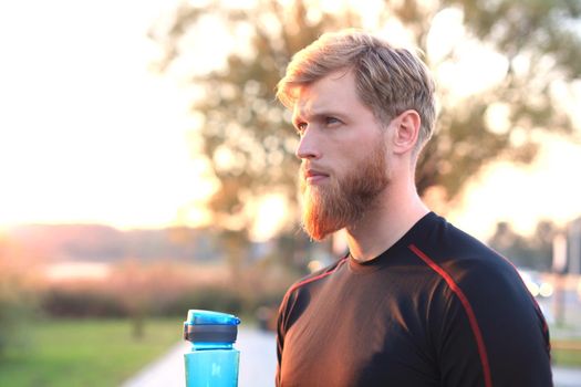 Handsome adult man drinking water from fitness bottle while standing outside, at sunset or sunrise. Runner