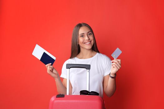Tourist girl in summer showing plastic credit card, with sunglasses, red suitcase, passport isolated on red background.
