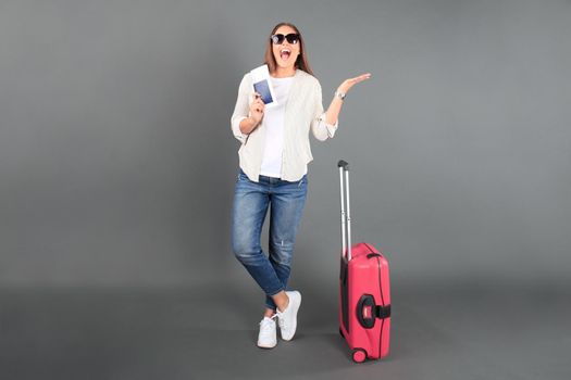 Young tourist girl in summer casual clothes, with sunglasses, red suitcase, passport, tickets isolated grey background.