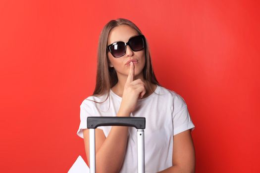 Young tourist girl in summer casual clothes, with sunglasses, red suitcase, passport isolated on red background.