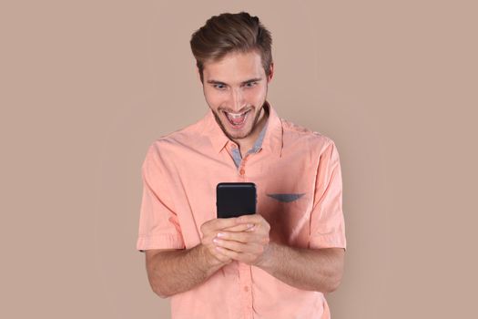 Shocked young man standing isolated over beige background, using mobile phone