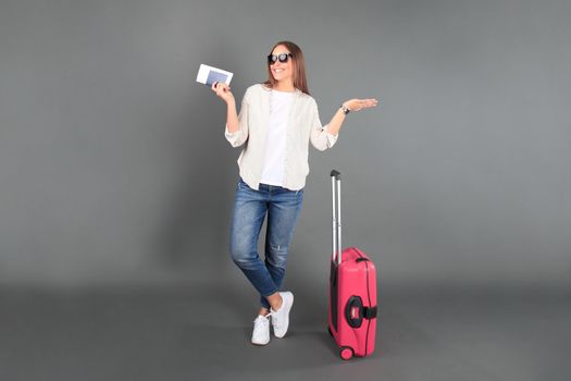 Young tourist girl in summer casual clothes, with sunglasses, red suitcase, passport, tickets isolated grey background.