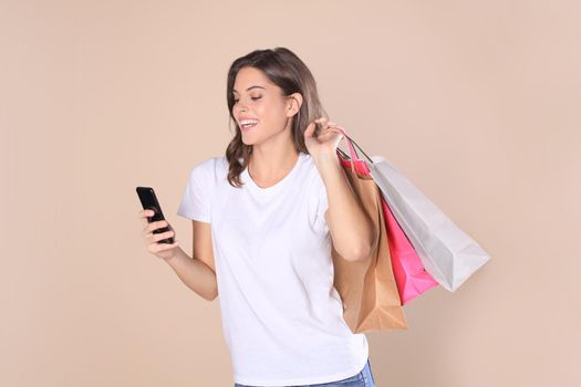 Cheerful young girl in basic clothes using mobile phone while holding shopping bags isolated over beige background