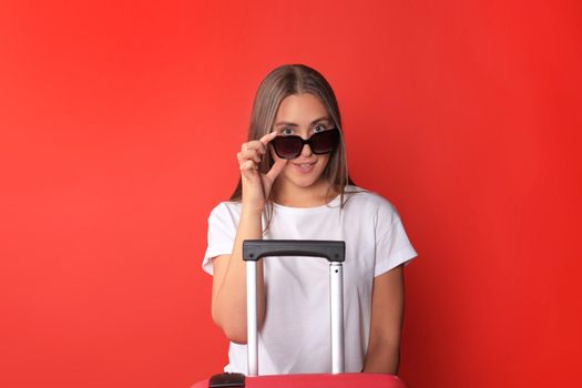 Young tourist girl in summer casual clothes, with sunglasses, red suitcase, passport isolated on red background.