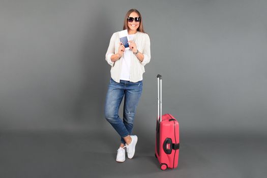 Young tourist girl in summer casual clothes, with sunglasses, red suitcase, passport, tickets isolated grey background.