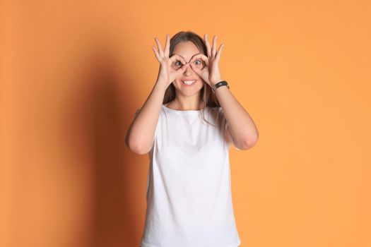 Young woman in basic clothing smiling looking at camera through holes made with fingers isolated over yellow background.