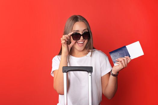 Young tourist girl in summer casual clothes, with sunglasses, red suitcase, passport isolated on red background.