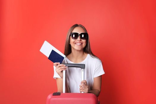 Young tourist girl in summer casual clothes, with sunglasses, red suitcase, passport isolated on red background.