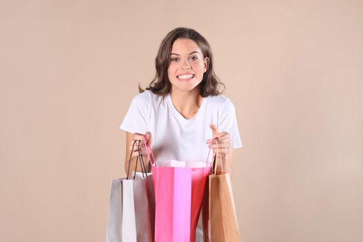 Cheerful young girl in basic clothes using mobile phone while holding shopping bags isolated over beige background