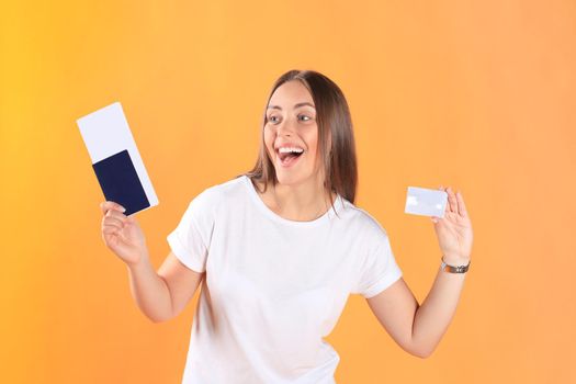 Excited young woman tourist standing isolated on yellow background holding passport with tickets, plastic credit card.