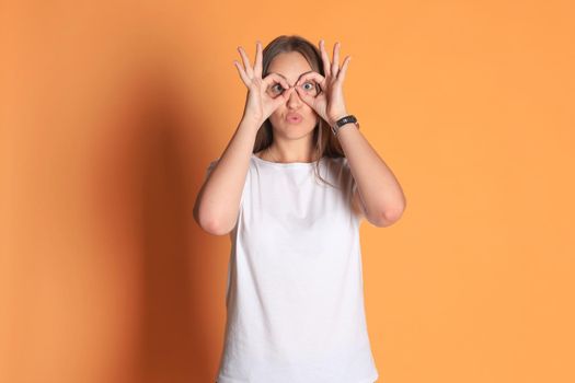Young woman in basic clothing smiling looking at camera through holes made with fingers isolated over yellow background.