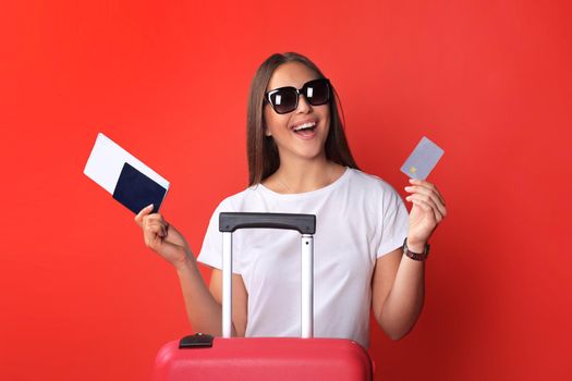 Young tourist girl in summer casual clothes, with sunglasses, red suitcase, passport isolated on red background.