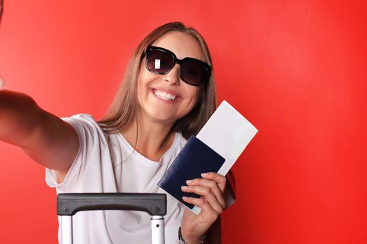 Young tourist girl in summer casual clothes, with sunglasses, red suitcase, passport isolated on red background.