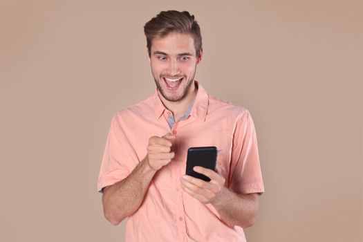 Shocked young man standing isolated over beige background, using mobile phone