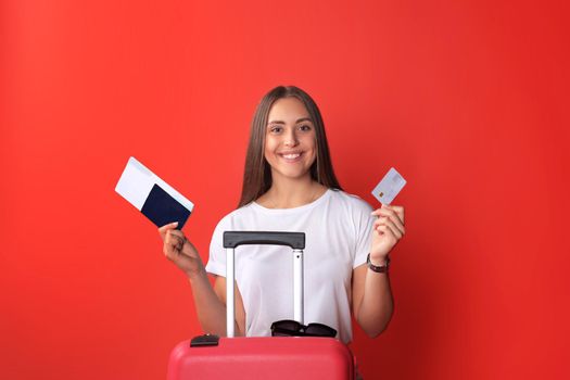 Young tourist girl in summer casual clothes, with sunglasses, red suitcase, passport isolated on red background.