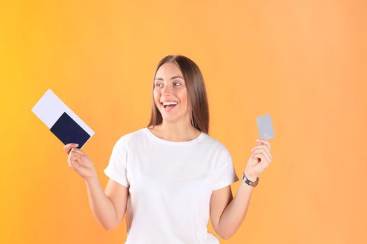 Excited young woman tourist standing isolated on yellow background holding passport with tickets, plastic credit card.