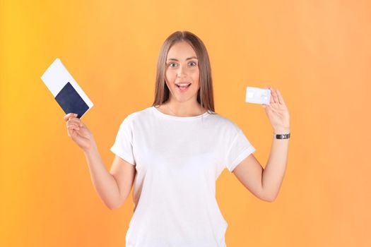 Excited young woman tourist standing isolated on yellow background holding passport with tickets, plastic credit card.