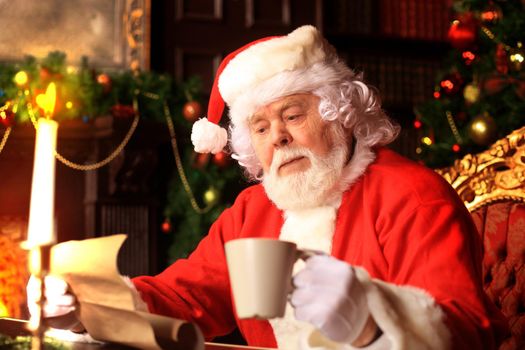 Portrait of happy Santa Claus sitting at his room at home near Christmas tree and reading Christmas letter or wish list.