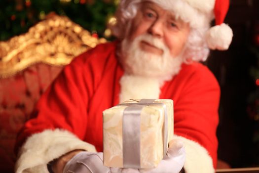 Portrait of happy Santa Claus sitting at his room at home near Christmas tree with gift box.