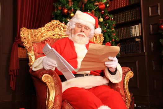 Portrait of happy Santa Claus sitting at his room at home near Christmas tree and reading Christmas letter or wish list.