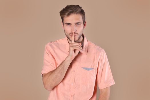 Young man with bristle posing on camera and holding index finger on lips isolated over beige background