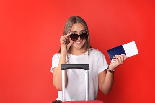 Young tourist girl in summer casual clothes, with sunglasses, red suitcase, passport isolated on red background.