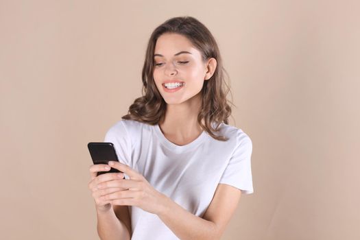 Young woman dressed in basic clothing isolated on beige background, using mobile phone