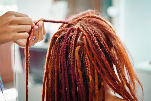 Stylish therapy professional care concept. Close up of braiding process plait with colored kanekalon. Hippie style hairstyle. Beauty salon services. Woman hairdresser weaves girl ginger dreadlocks.