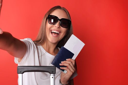 Young tourist girl in summer casual clothes, with sunglasses, red suitcase, passport isolated on red background.