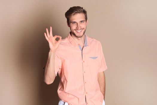 Smiling young man standing isolated over beige background, showing ok gesture