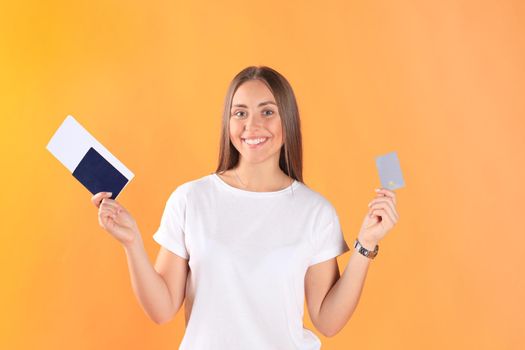 Excited young woman tourist standing isolated on yellow background holding passport with tickets, plastic credit card.